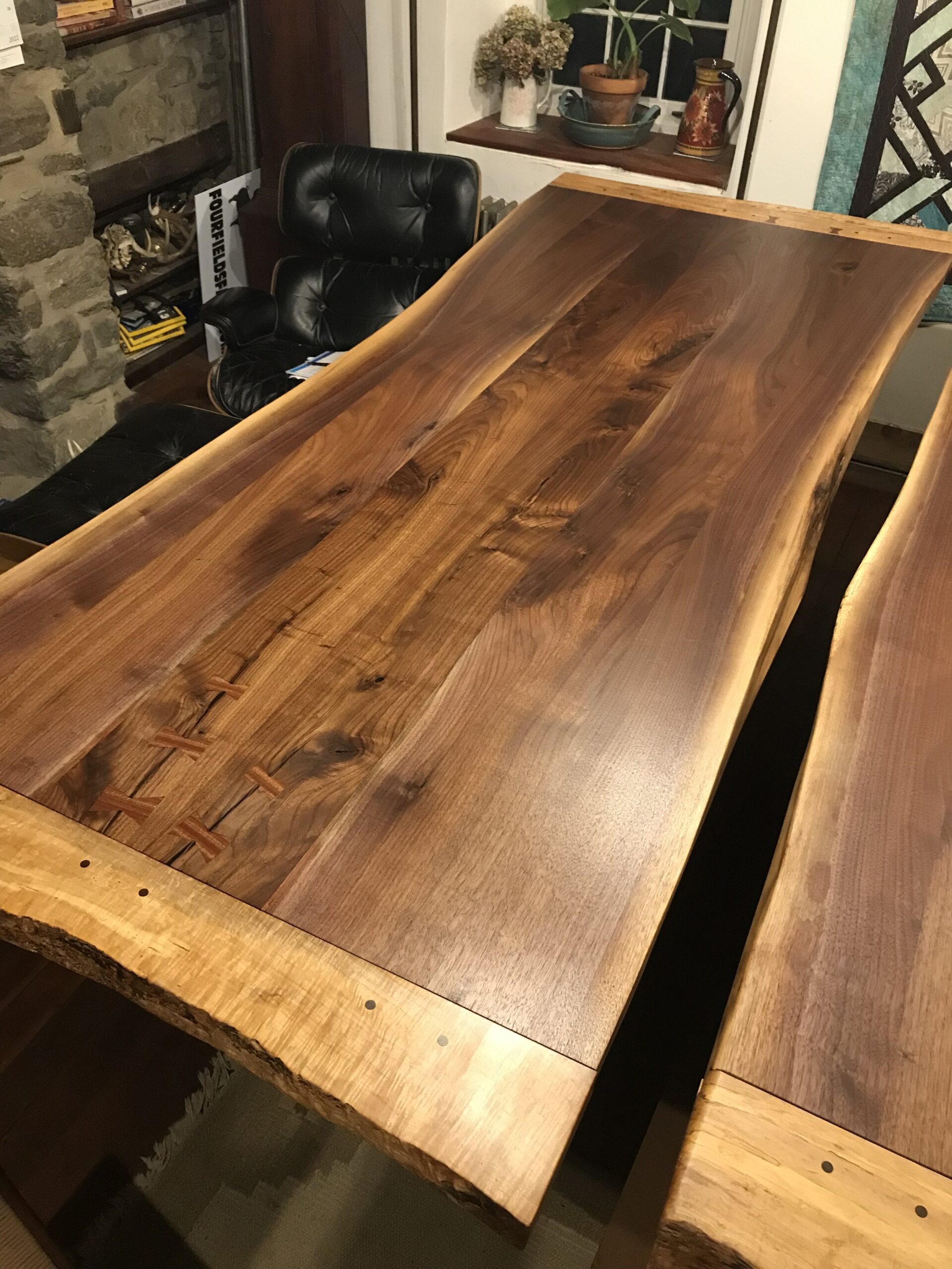 IMG_5945 Close-up of a handcrafted wooden table showcasing rich walnut grain and live edge detailing, with visible joinery and contrasting lighter wood accents. The table is set in a cozy interior space featuring a stone wall and black leather chair.