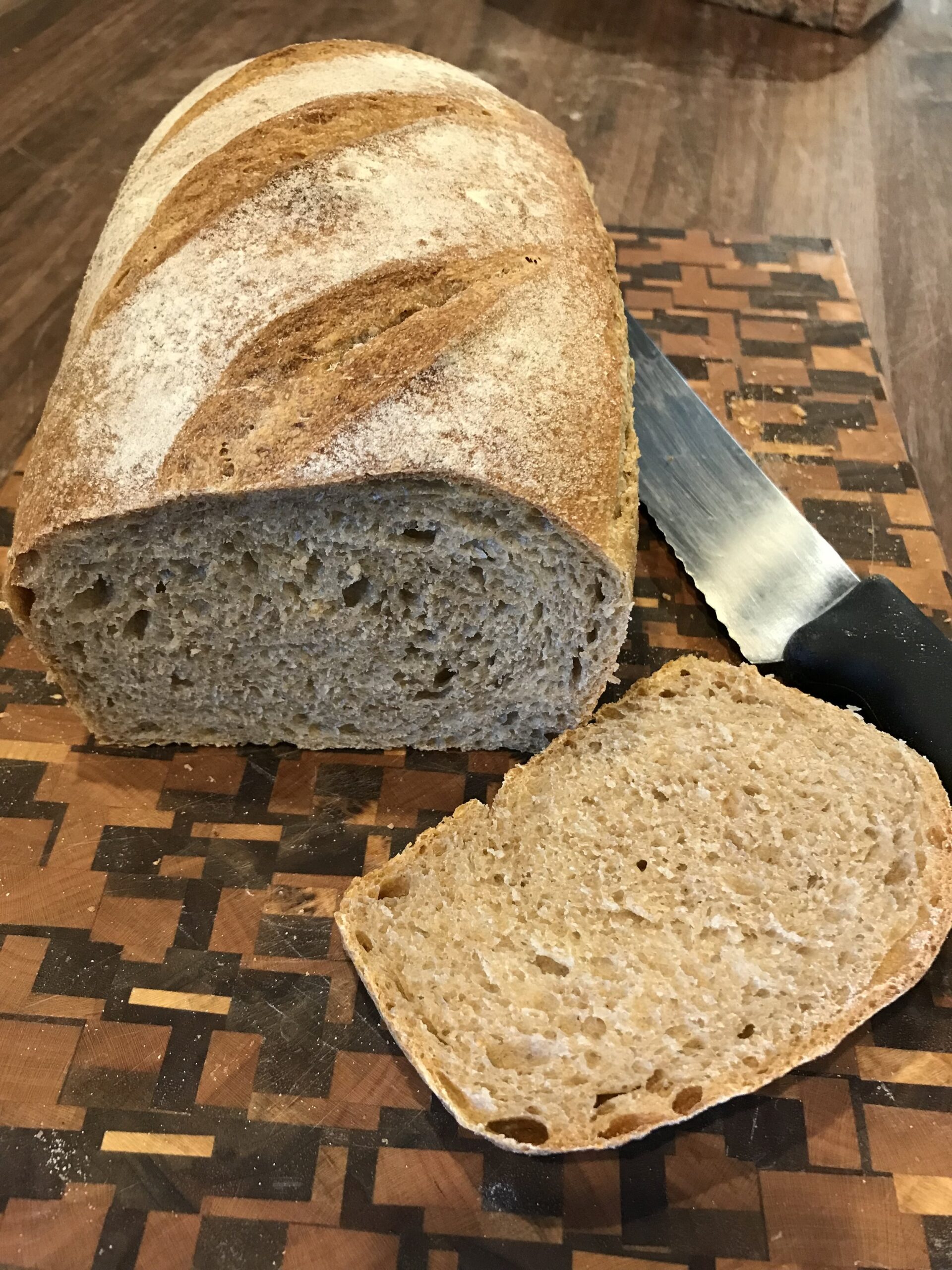 IMG_6987 Freshly baked whole grain bread loaf on a wooden cutting board, with a slice cut off and a knife placed beside it. The bread features a crusty exterior with distinctive slashes on top, showcasing its artisanal quality.