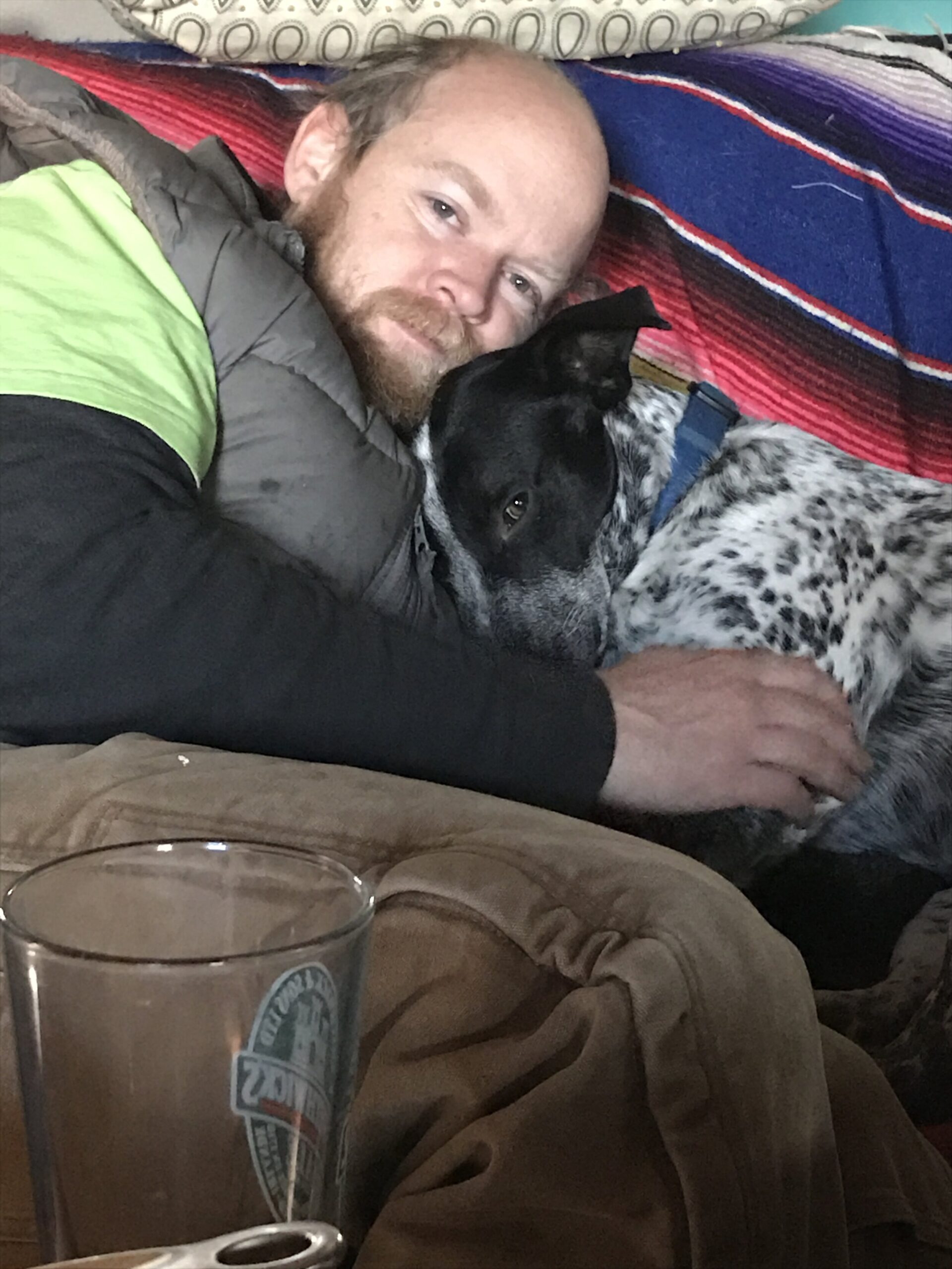 Liz+Balchin+-+IMG_3572 A man cuddles with a black and white dog on a cozy couch, showcasing a warm moment of companionship. A glass is visible in the foreground, adding to the relaxed atmosphere of the scene.