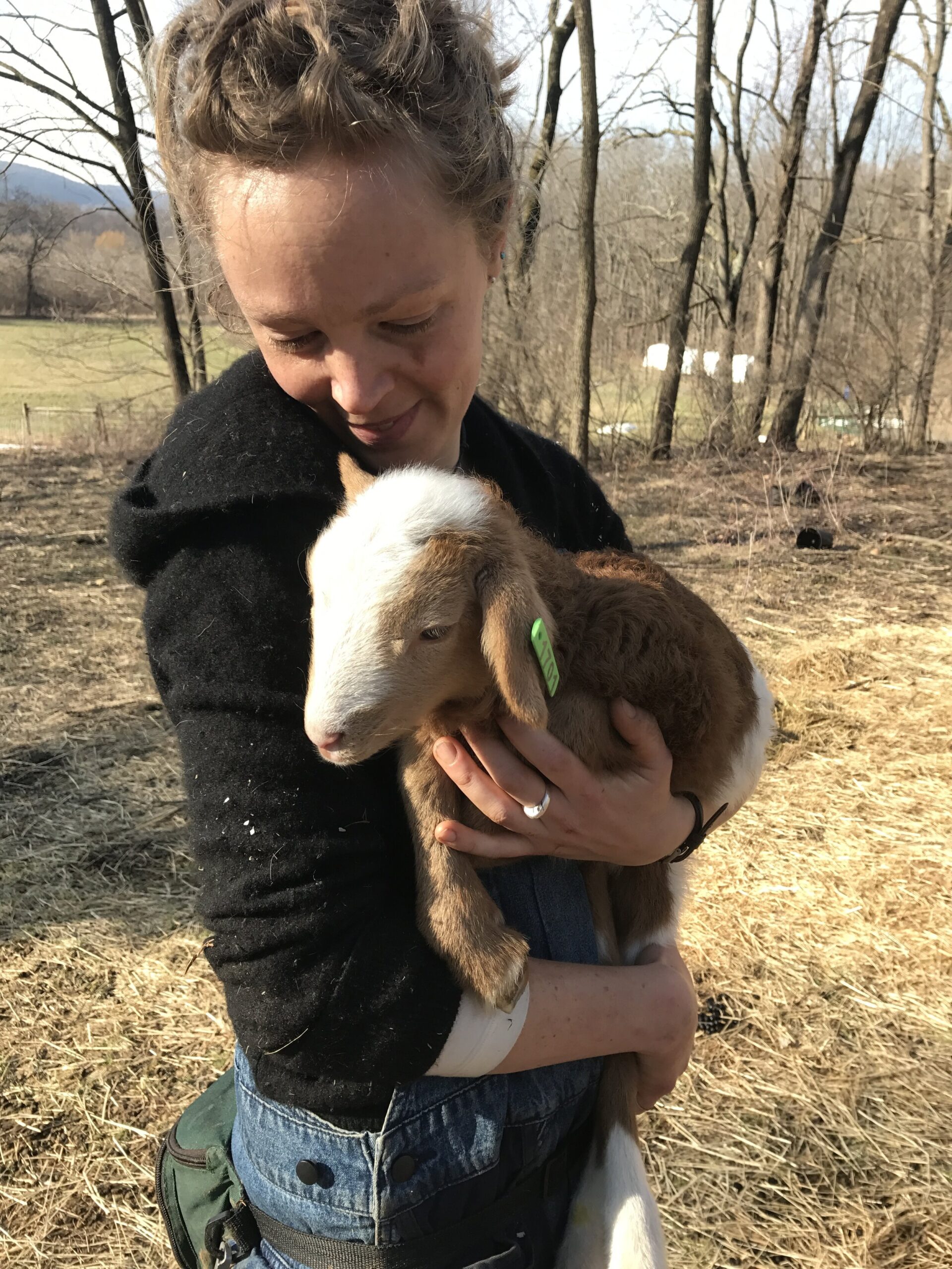 Liz+Balchin+-+IMG_4138 Woman holding a young goat in a pastoral setting, surrounded by trees and fields, showcasing a connection between humans and farm animals.